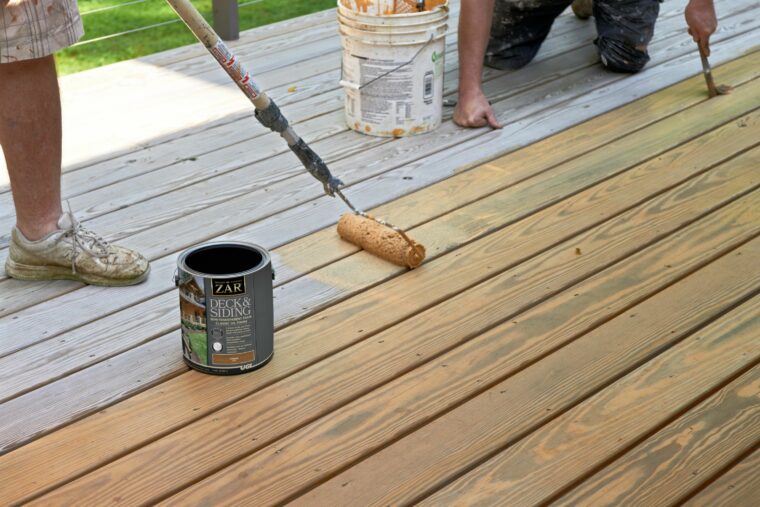 Worker applying deck coating with a roller on a wood deck beside a can of ZAR Deck and Siding paint